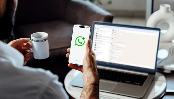 An undated image shows person holding a cup of tea with smartphone, on which WhatsApp logo is displayed and WhatsApp Web on the laptop in background. — Shutterstock